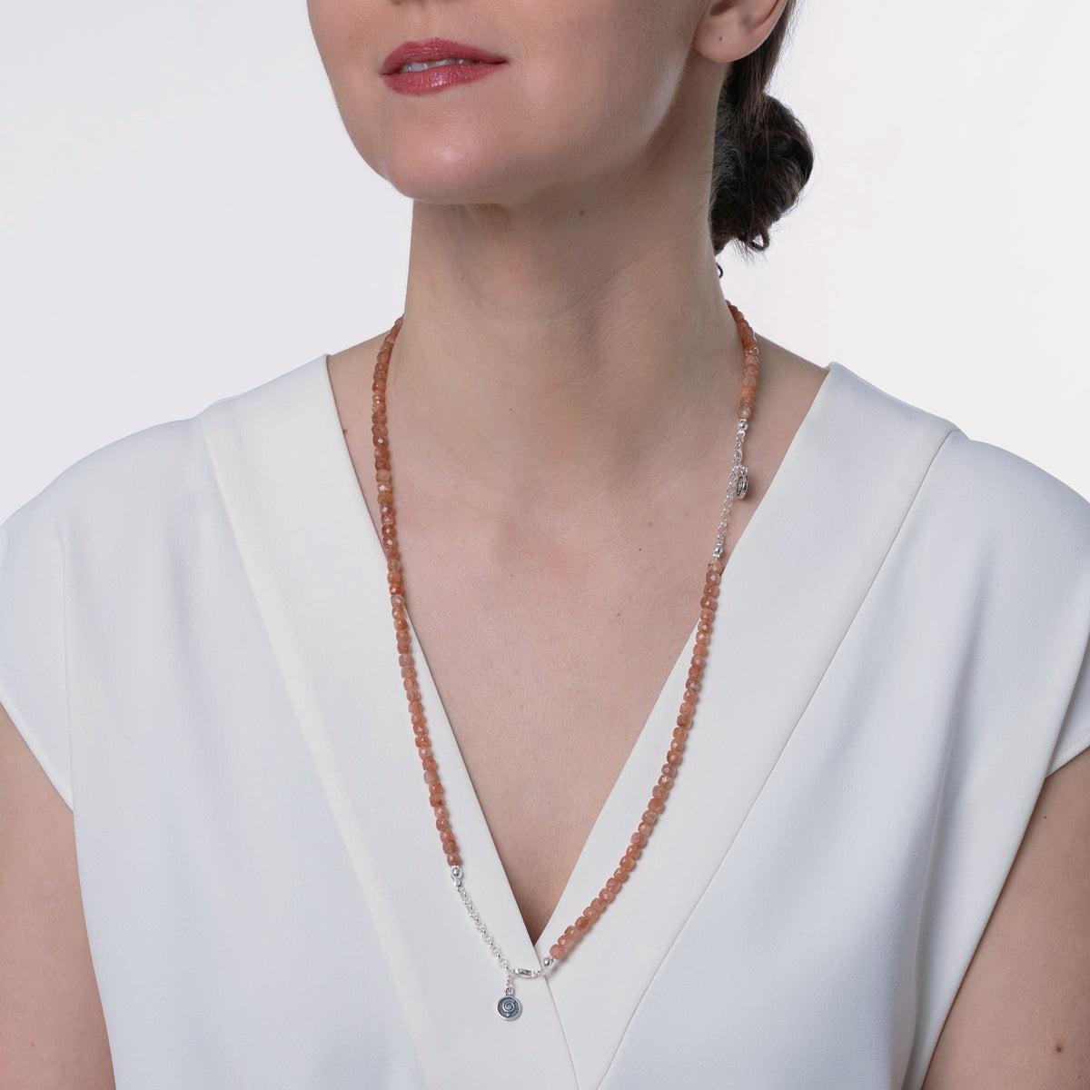A close-up of a woman wearing a long beaded necklace with peach-orange beads, a silver chain, and a small silver pendant, paired with a white V-neck top.